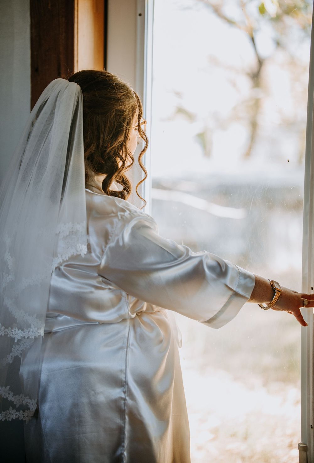 A bride in a white silk robe and veil looks out a window on her wedding morning with soft natural light.