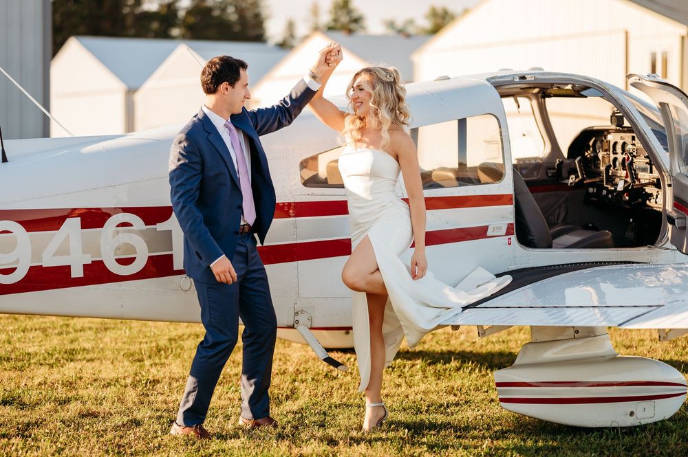 Two people in wedding attire share a celebratory high-five in front of a small private aircraft on a grassy airfield.