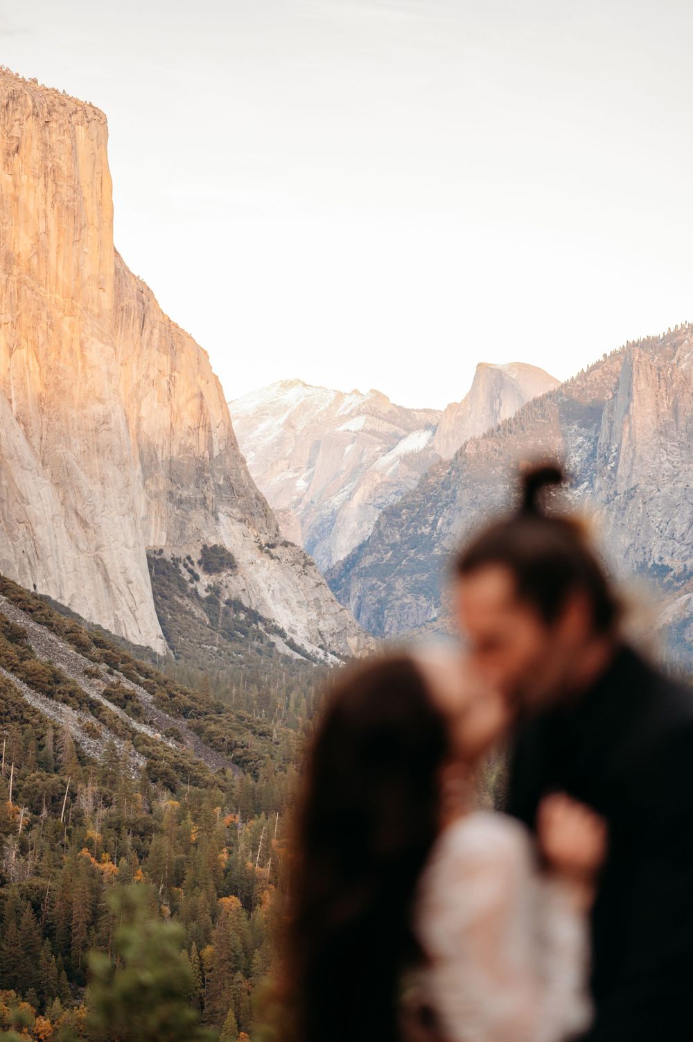 Romantic couple embraces at scenic Yosemite National Park overlook with El Capitan and valley cliffs glowing at sunset.