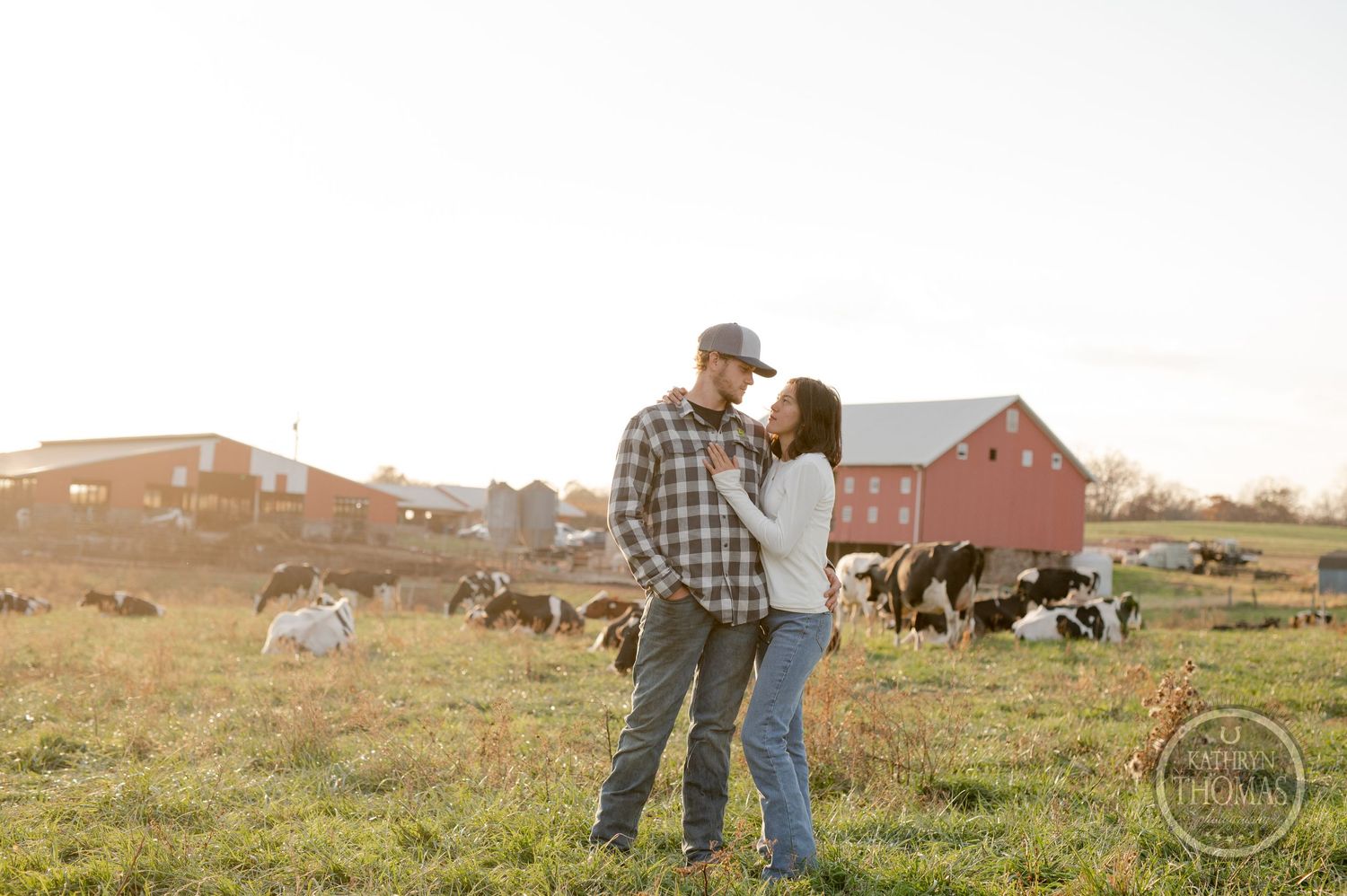 Two people share an intimate moment while standing in a pastoral field with barns and cows in the background.