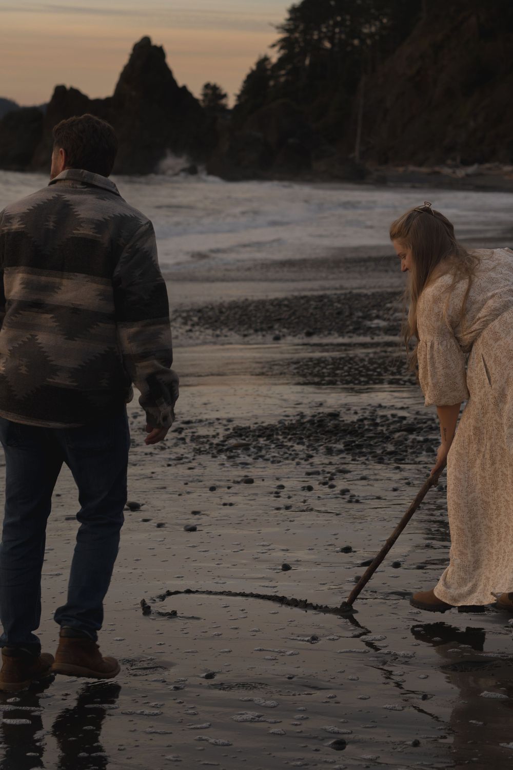 Couple drawing in sand on moody beach at sunset near rocky cliffs.