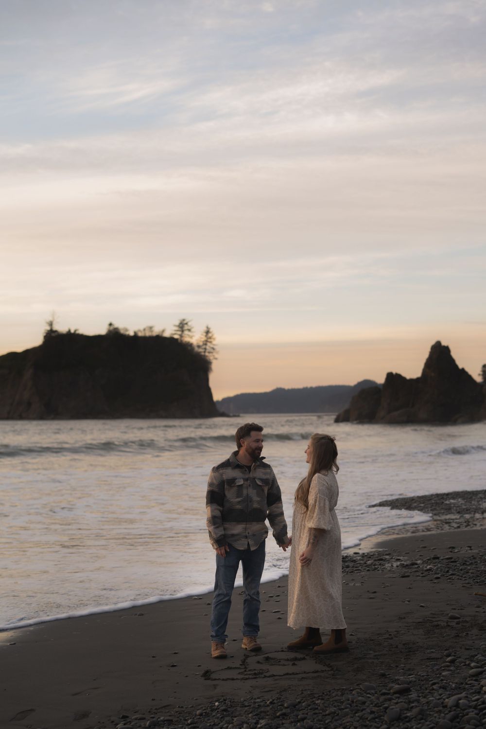 A couple shares a romantic moment at sunset on a rocky beach with dramatic coastal cliffs in the background. on Ruby Beach, Olympic National