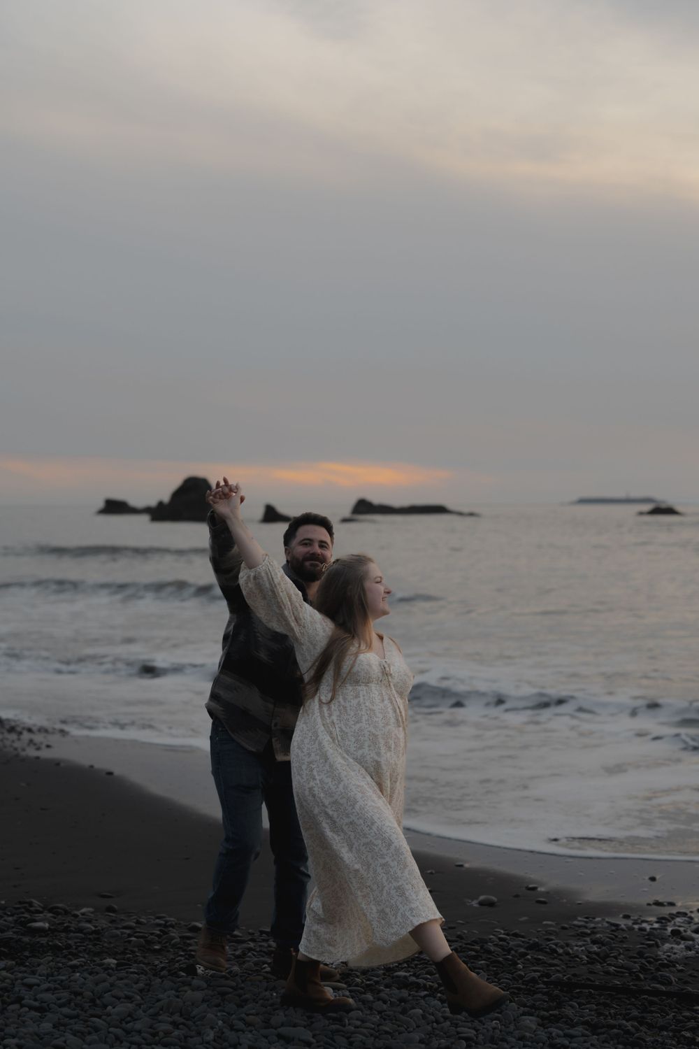Couple in romantic dance pose on beach at dusk with waves crashing behind them on Ruby Beach, Olympic National Park.