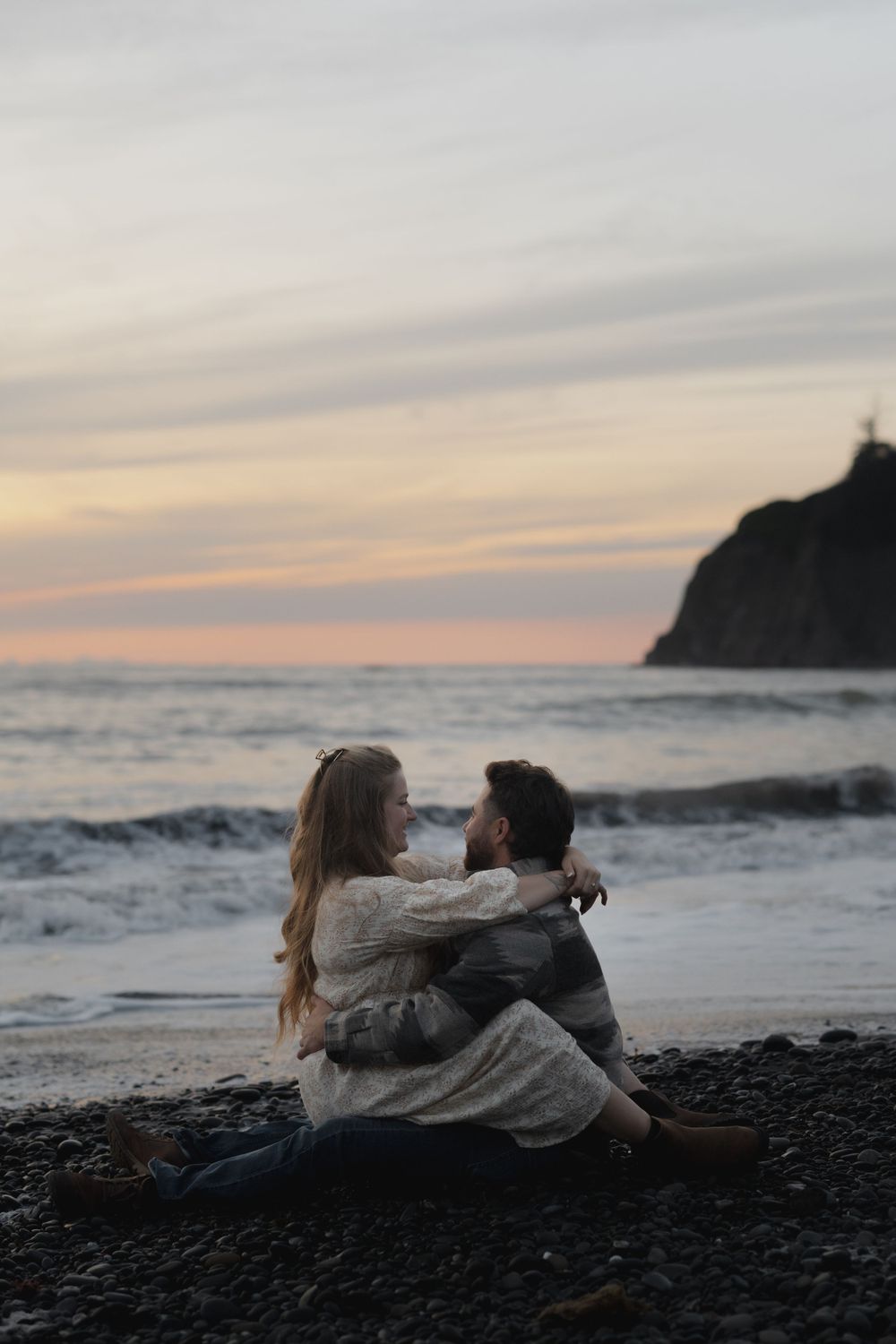 Couple embracing on black sand beach at sunset with dramatic coastal cliffs in background on Ruby Beach, Olympic National Park.