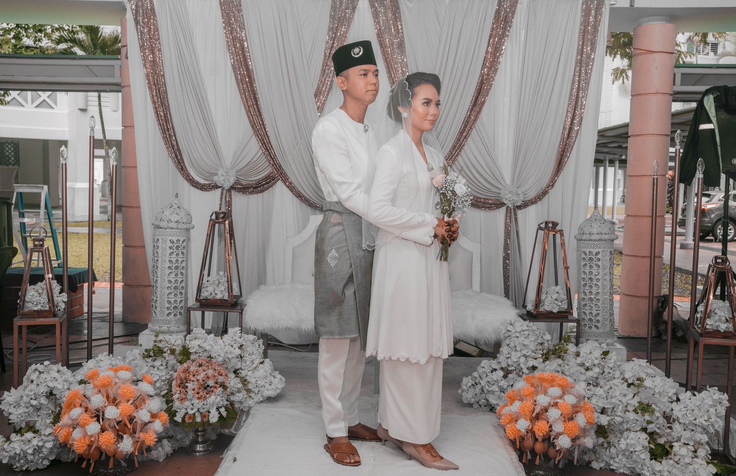 A couple stands together in traditional Malay wedding attire with orange floral decorations in an elegantly draped venue.