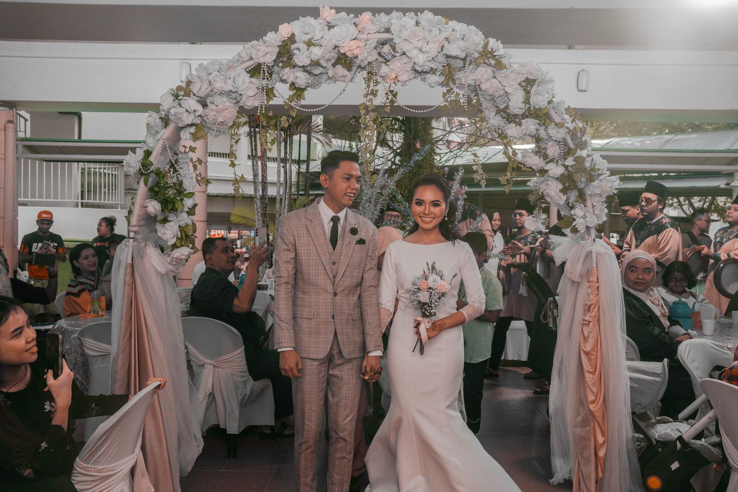 A couple poses under a floral arch decorated with pink and white flowers at their wedding ceremony.