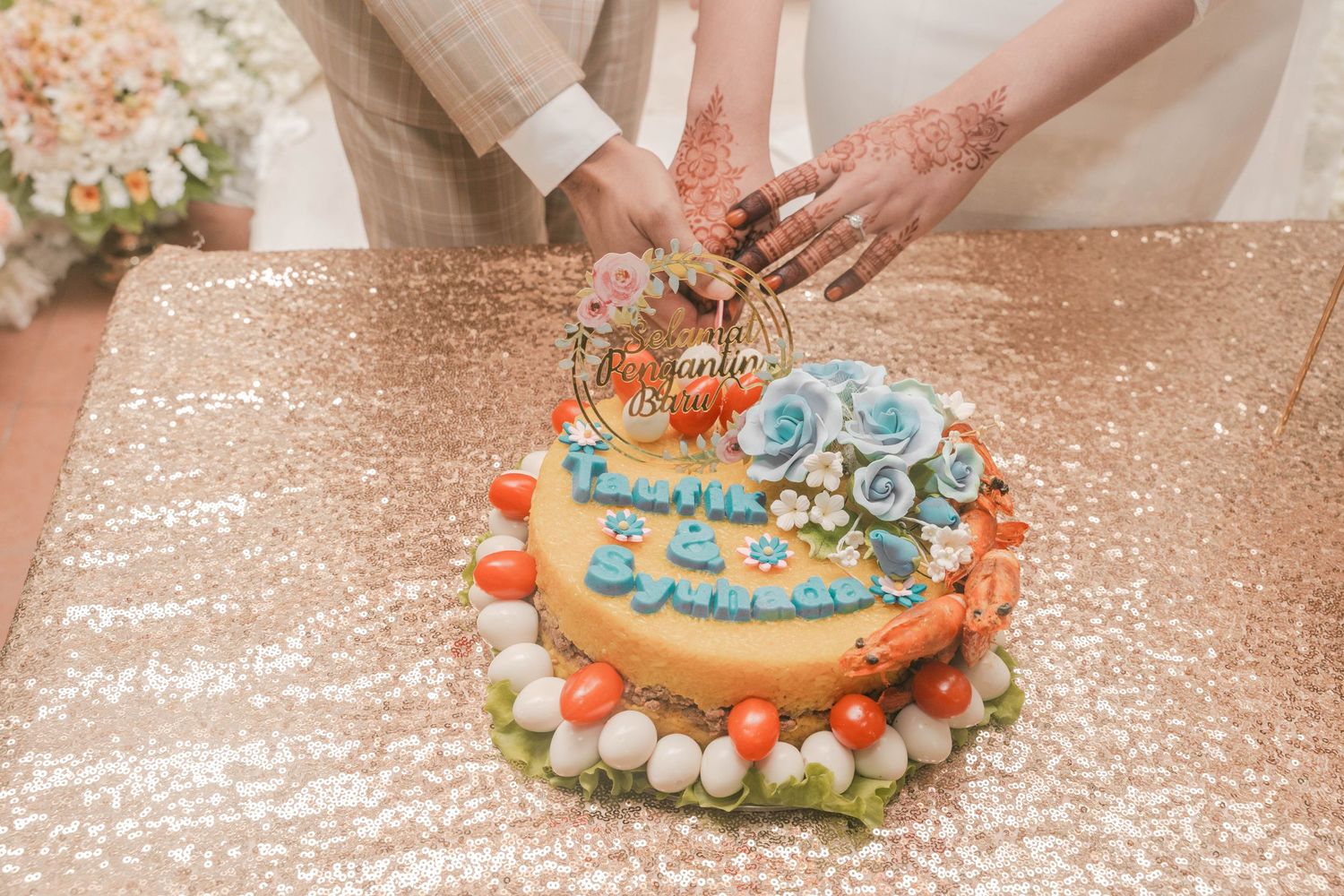 Hands cutting into an ornate wedding cake decorated with blue roses and pearls on a sparkly tablecloth.