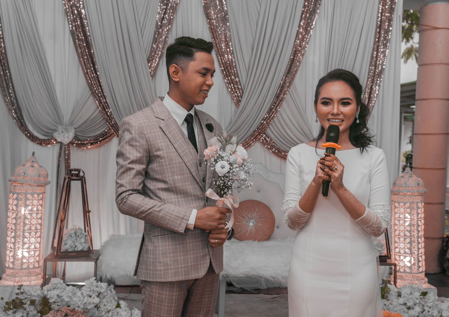 A couple holds microphones while speaking during their wedding ceremony against elegant draped decorations.