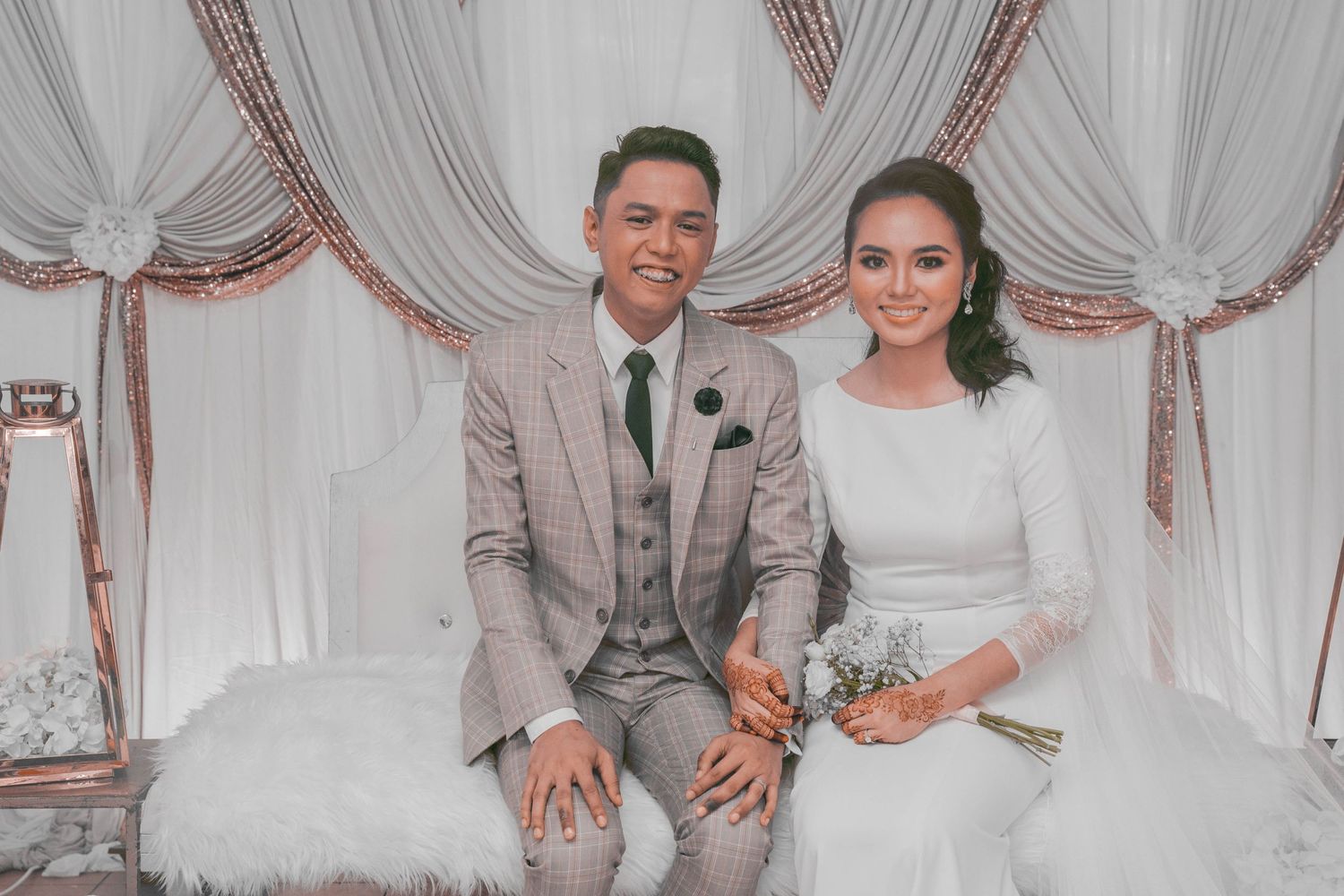 A couple sits together on an elegant white sofa against draped fabric wedding decorations.