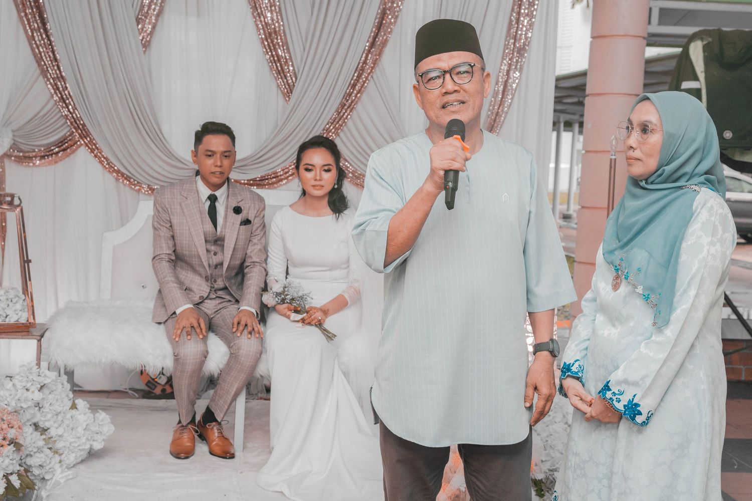 A speaker addresses guests at a wedding ceremony while the couple sits nearby.