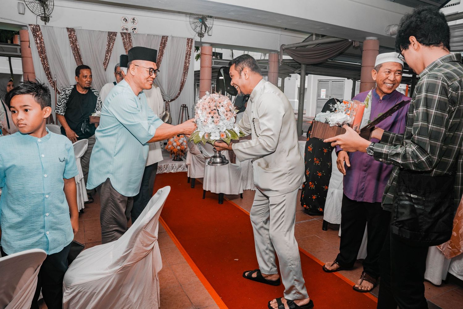 Traditional ceremony gathering with guests exchanging gifts on red carpet.