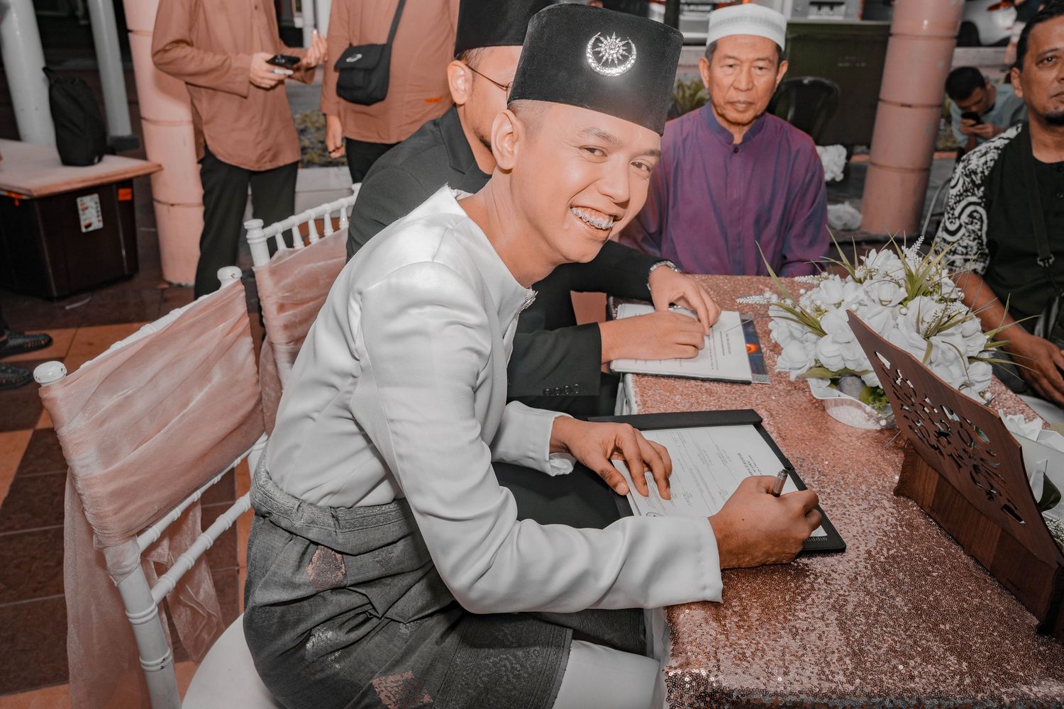 A signing ceremony at a wedding reception with guests watching from a decorated table.