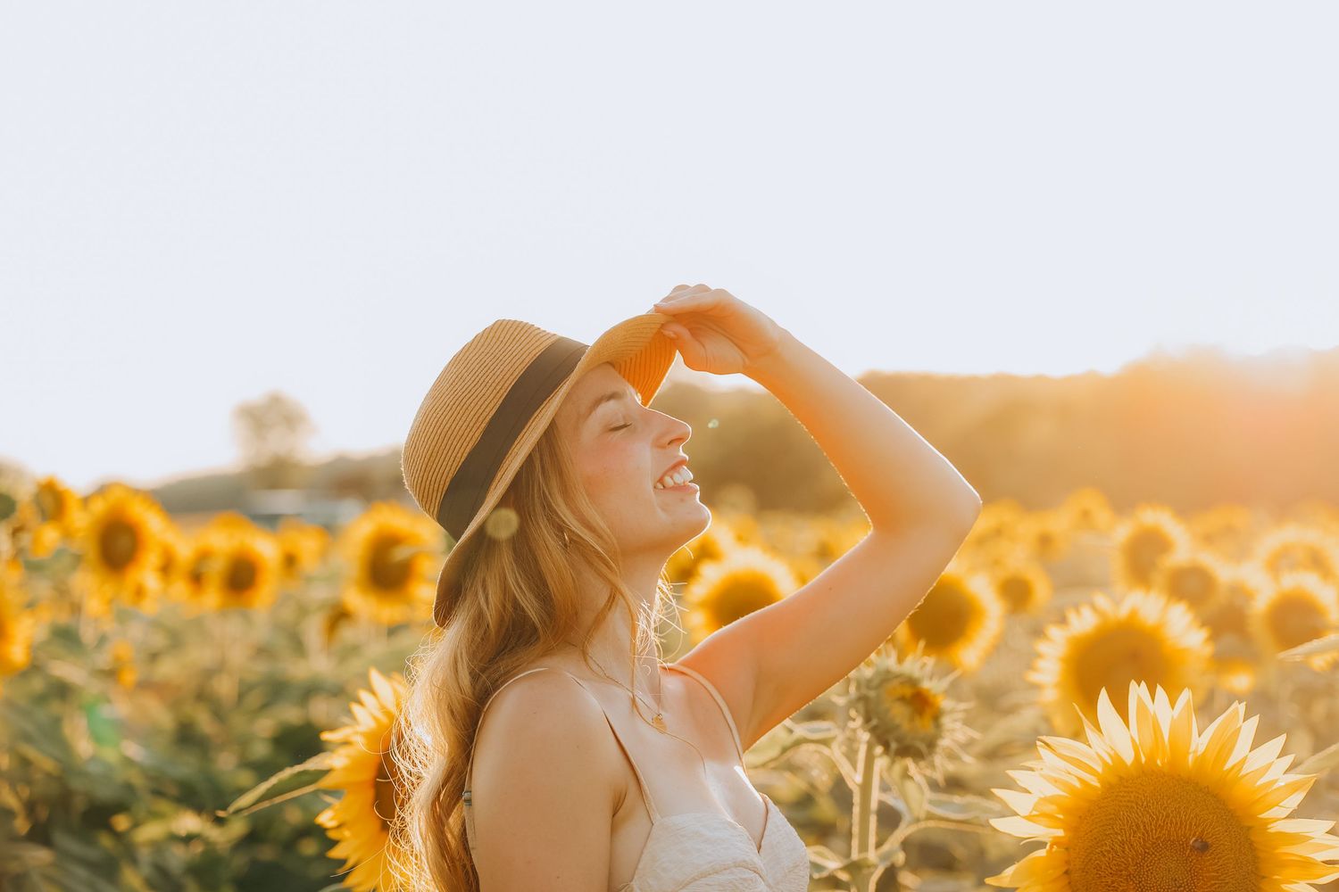 Woman in straw hat and white dress enjoying sunshine in a bright sunflower field.