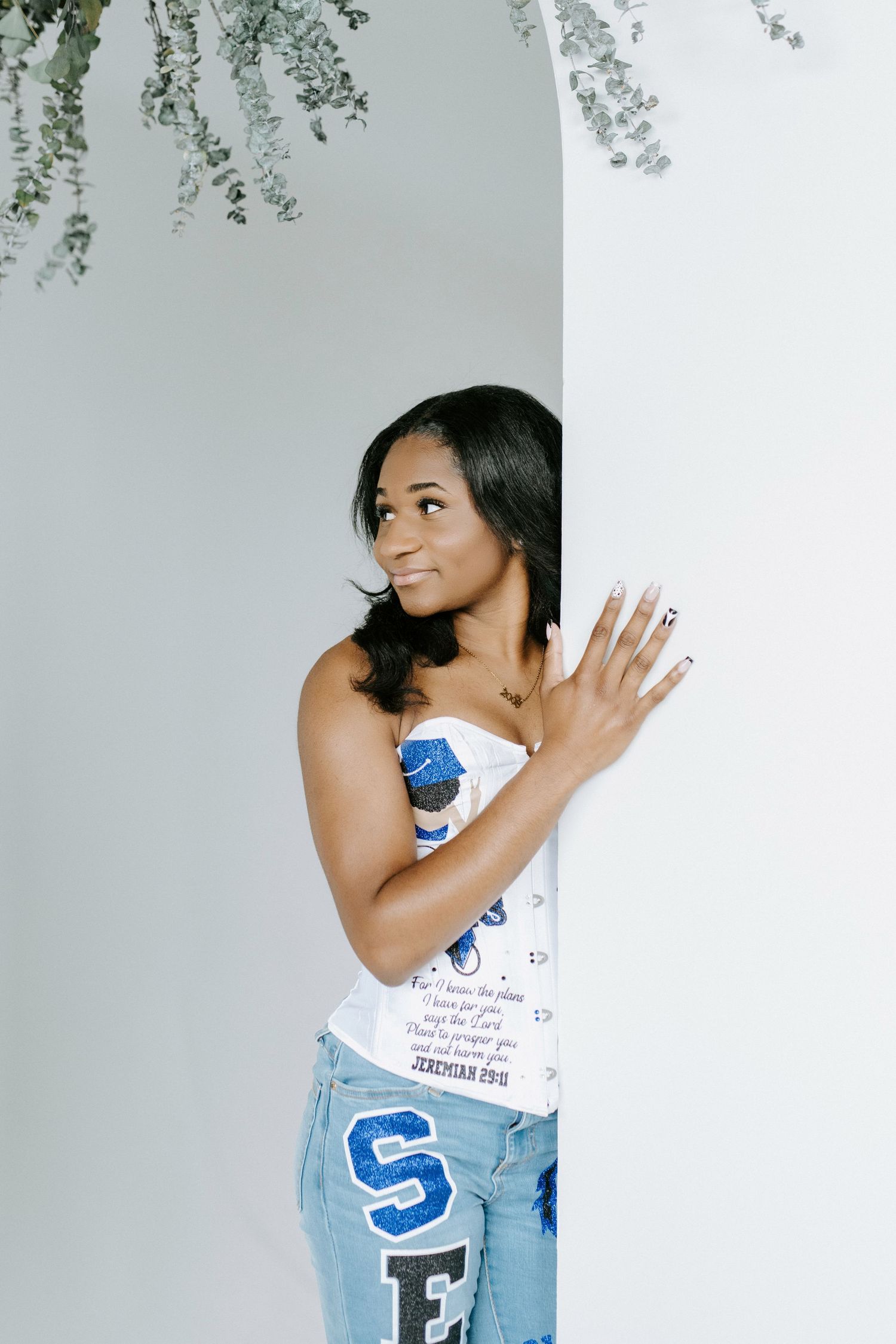 A person in a white tank top and blue jeans poses playfully against a light wall with foliage visible above.
