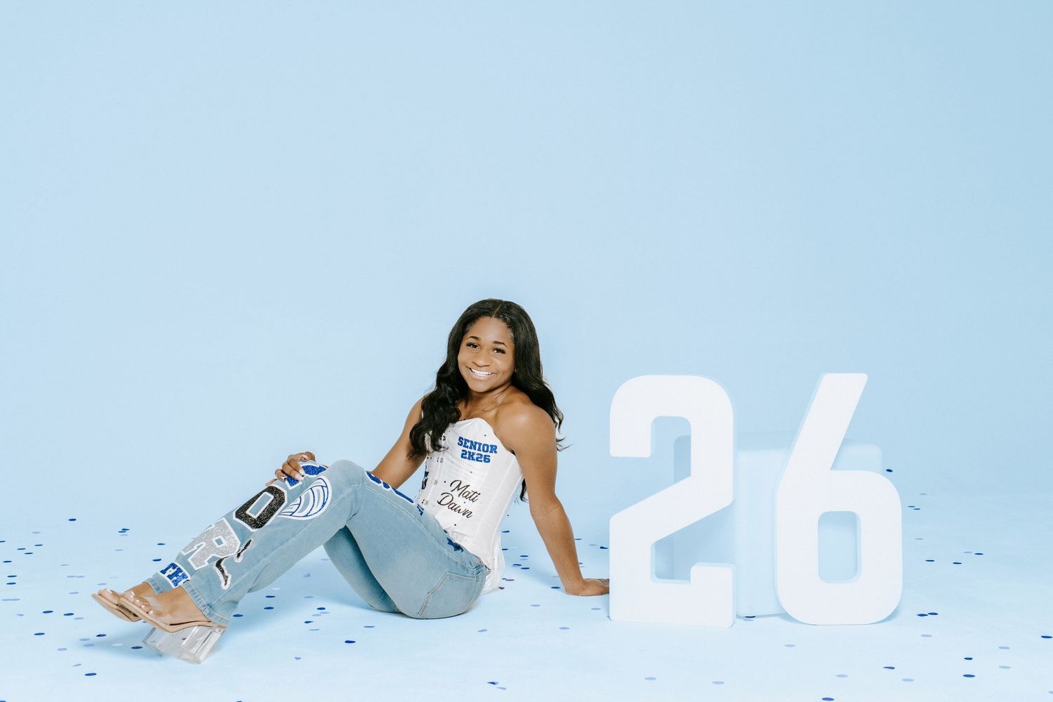 A casual senior portrait session features a sitting pose with large number 26 against a light blue background.