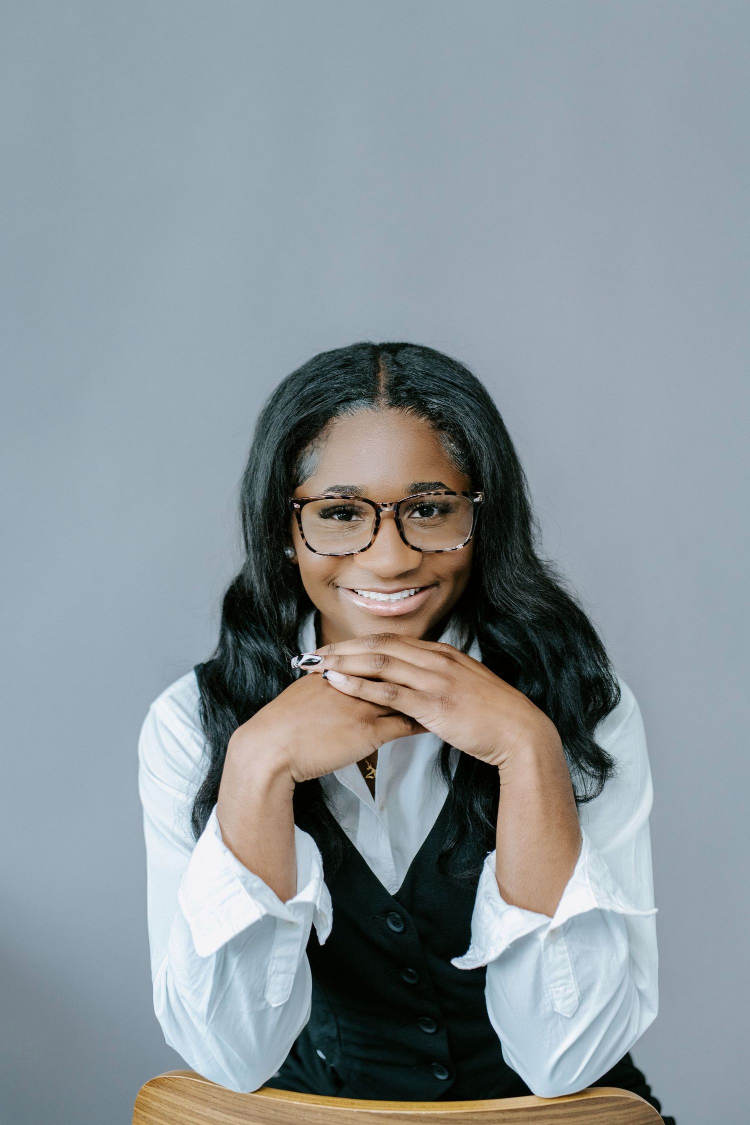 Professional portrait of someone in glasses, white button down shirt and black vest posing against grey backdrop with bright smile.