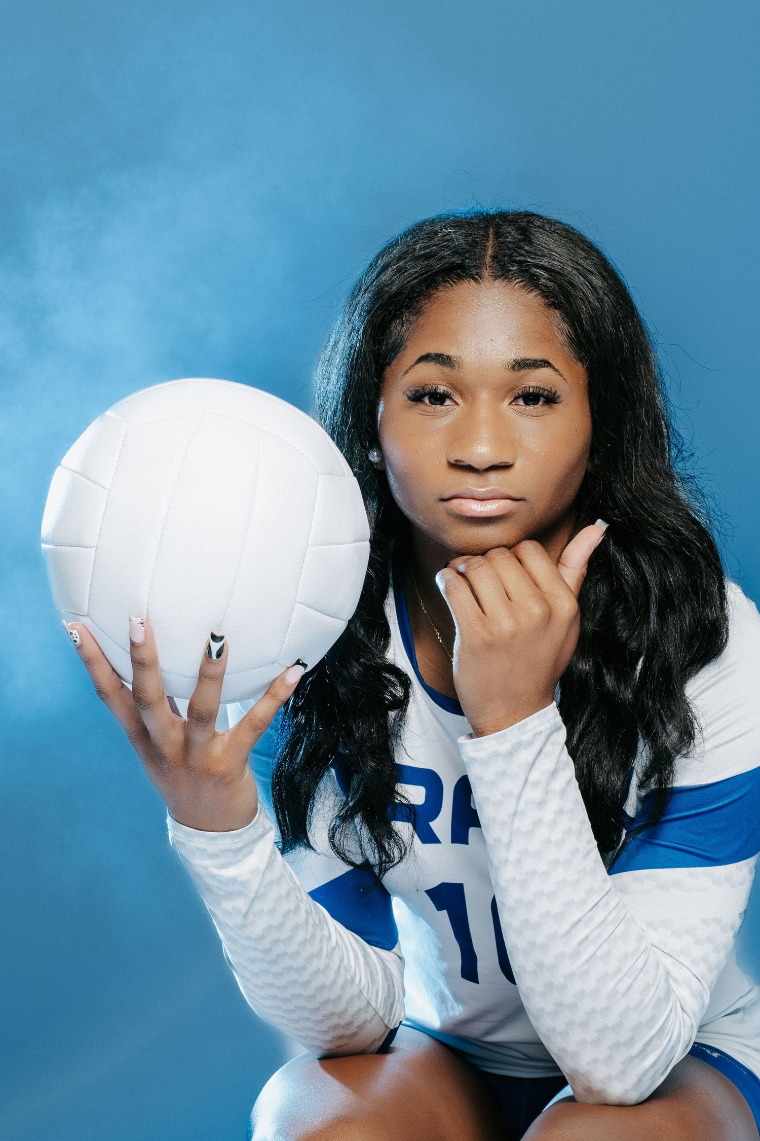 A person in a white sweater and blue jersey makes playful expressions while posing with a volleyball against a blue backdrop.