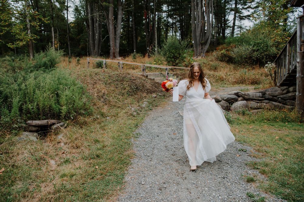 A bride in a white dress walks along a rocky forest path surrounded by tall evergreen trees and autumn foliage.