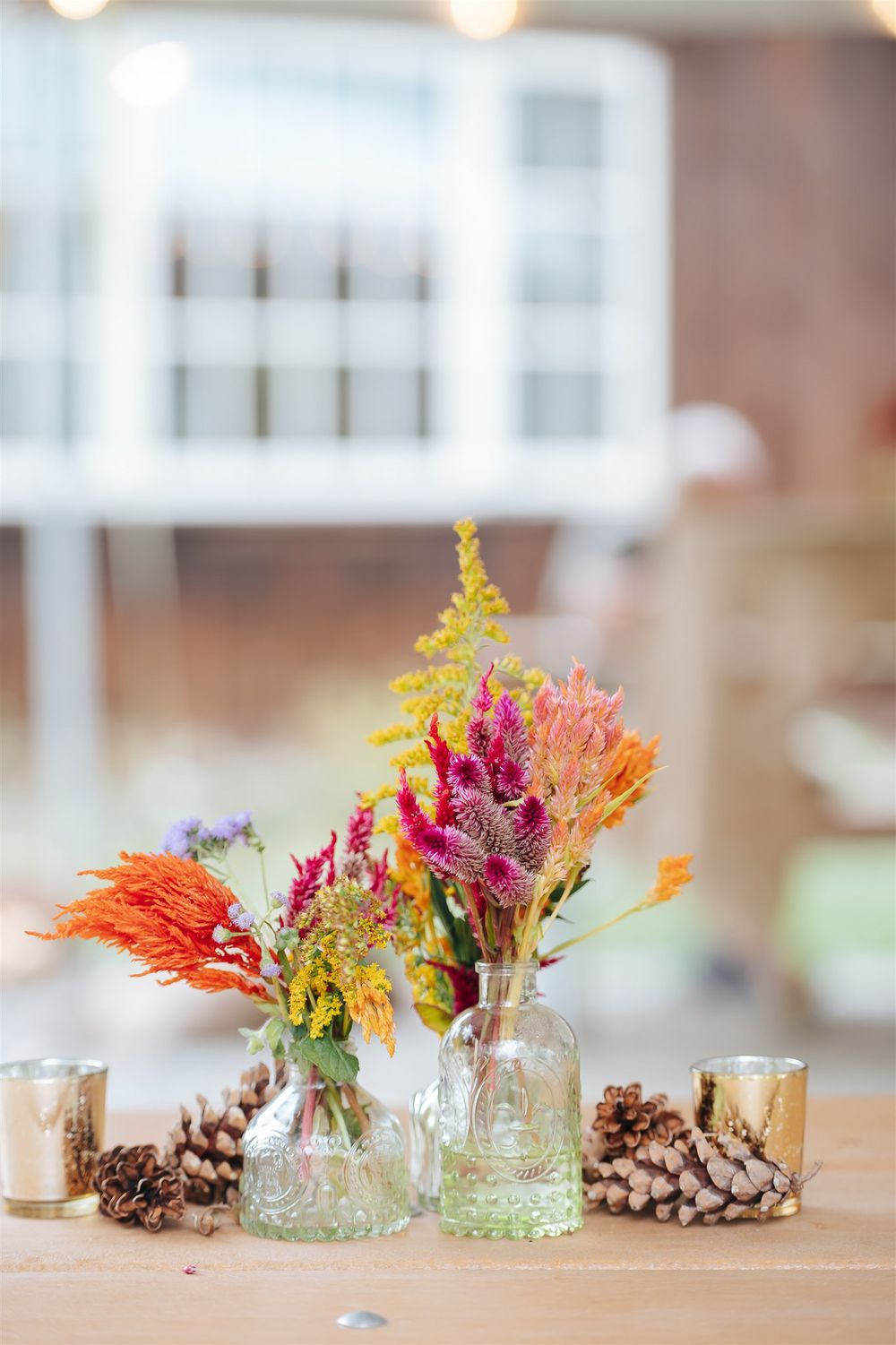 Glass vases filled with colorful autumn flowers and pinecones arranged as rustic wedding table centerpieces.