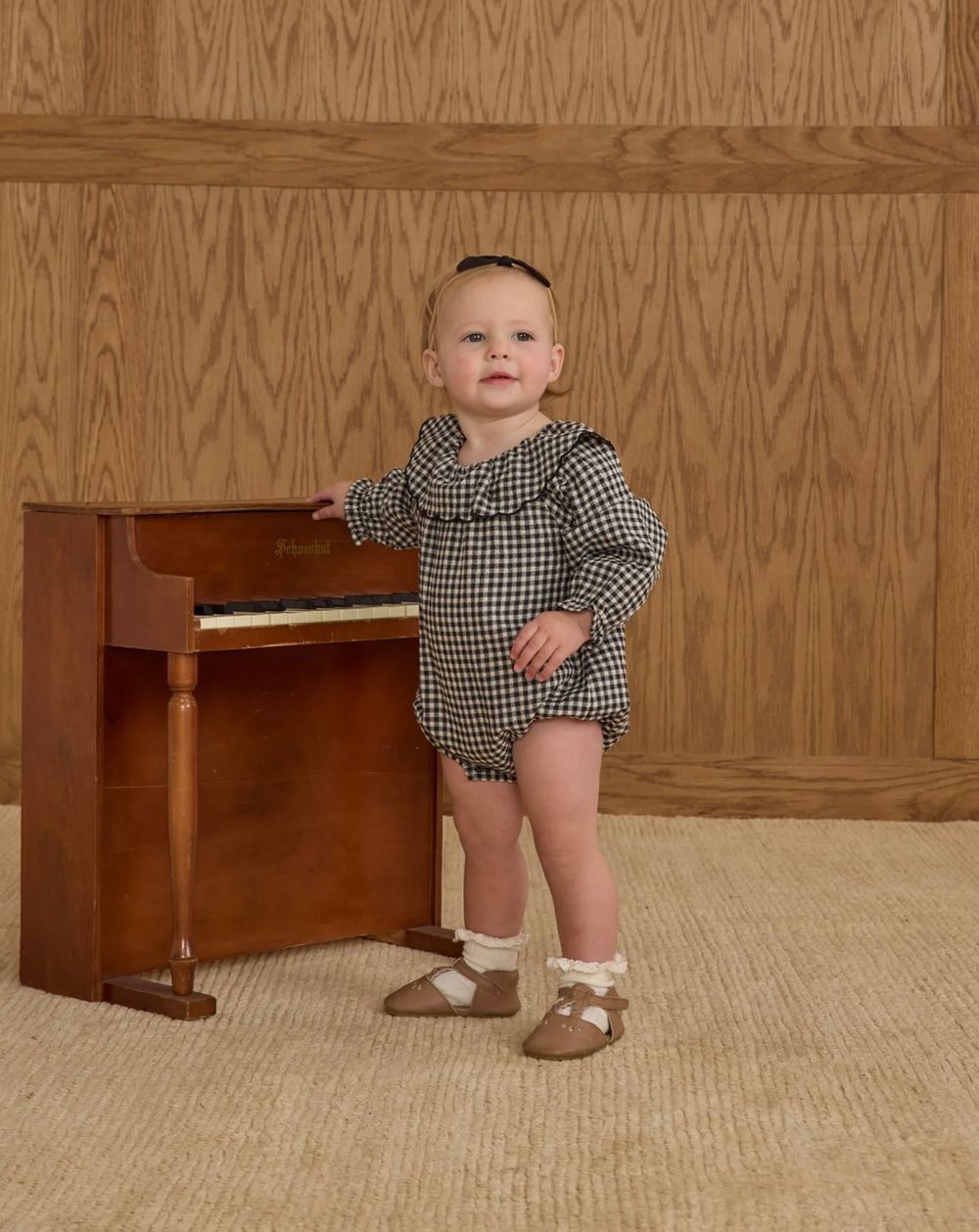 Small child in checkered outfit stands next to upright piano against wood-paneled wall.