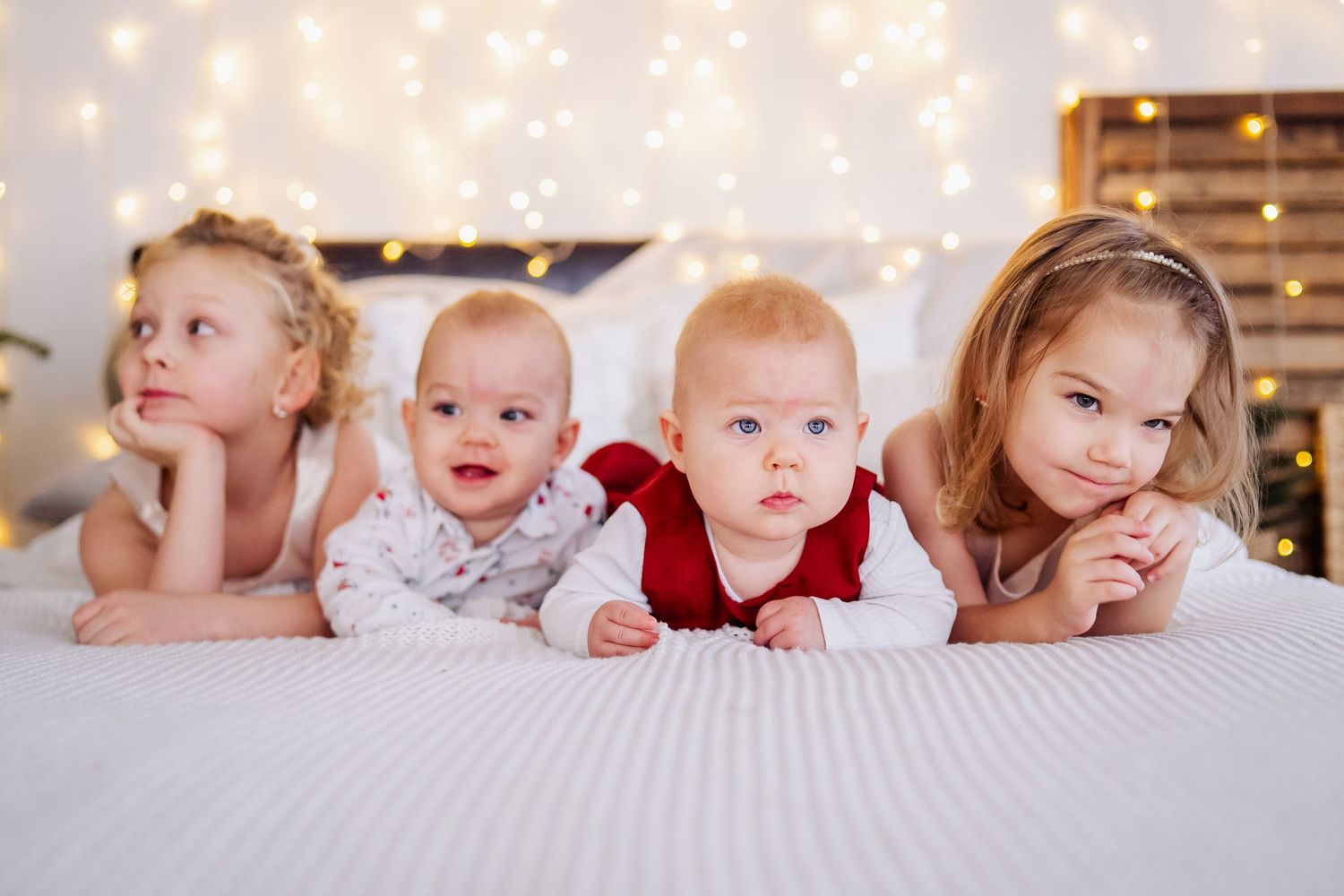 Four young children lying on a white bed with festive bokeh lights, including two babies and two toddler girls.
