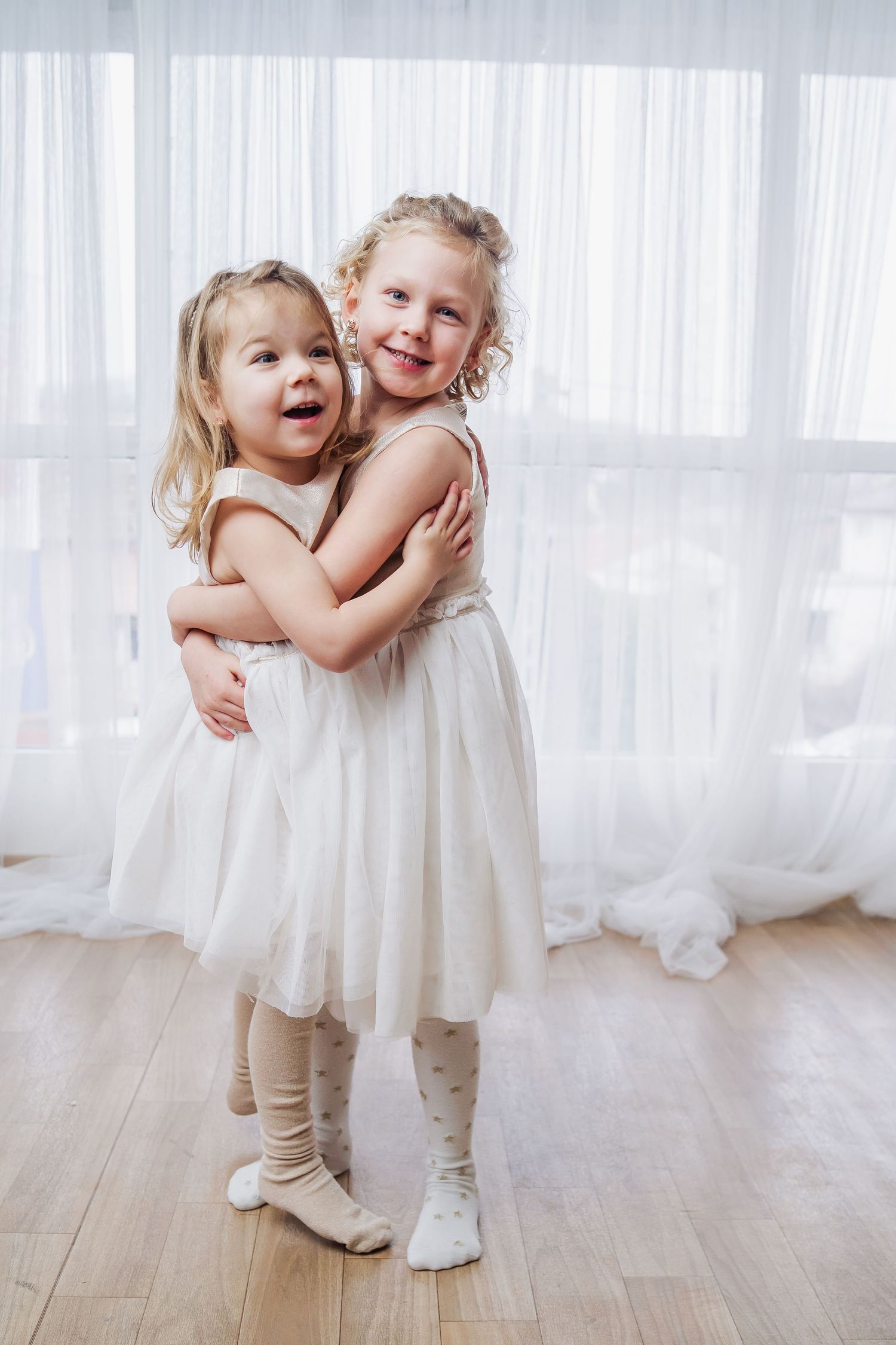 Two young girls in white tulle dresses hugging and smiling joyfully in a bright white room.