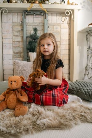 A young child in holiday plaid dress sits on a cozy gray blanket with teddy bears and decorative stone wall background.