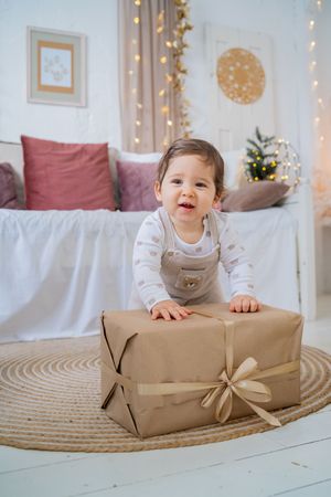 Sequence of photos showing a small child peeking over a large wrapped Christmas present on a round jute rug with twinkle lights.