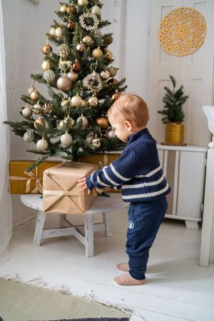 Small child in striped sweater reaches for wrapped present by decorated Christmas tree with gold and silver ornaments.