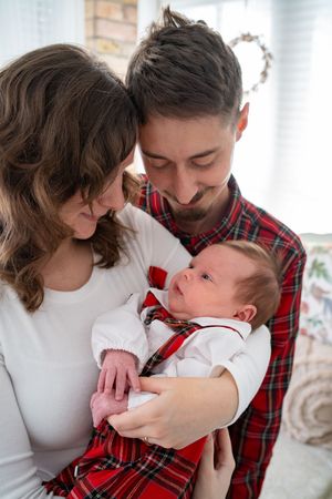 A couple in white sweater and red plaid shirt share tender moments with their newborn baby in heartwarming portraits.