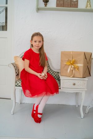 A child in a festive red party dress sits on a decorative white bench near wrapped holiday presents.
