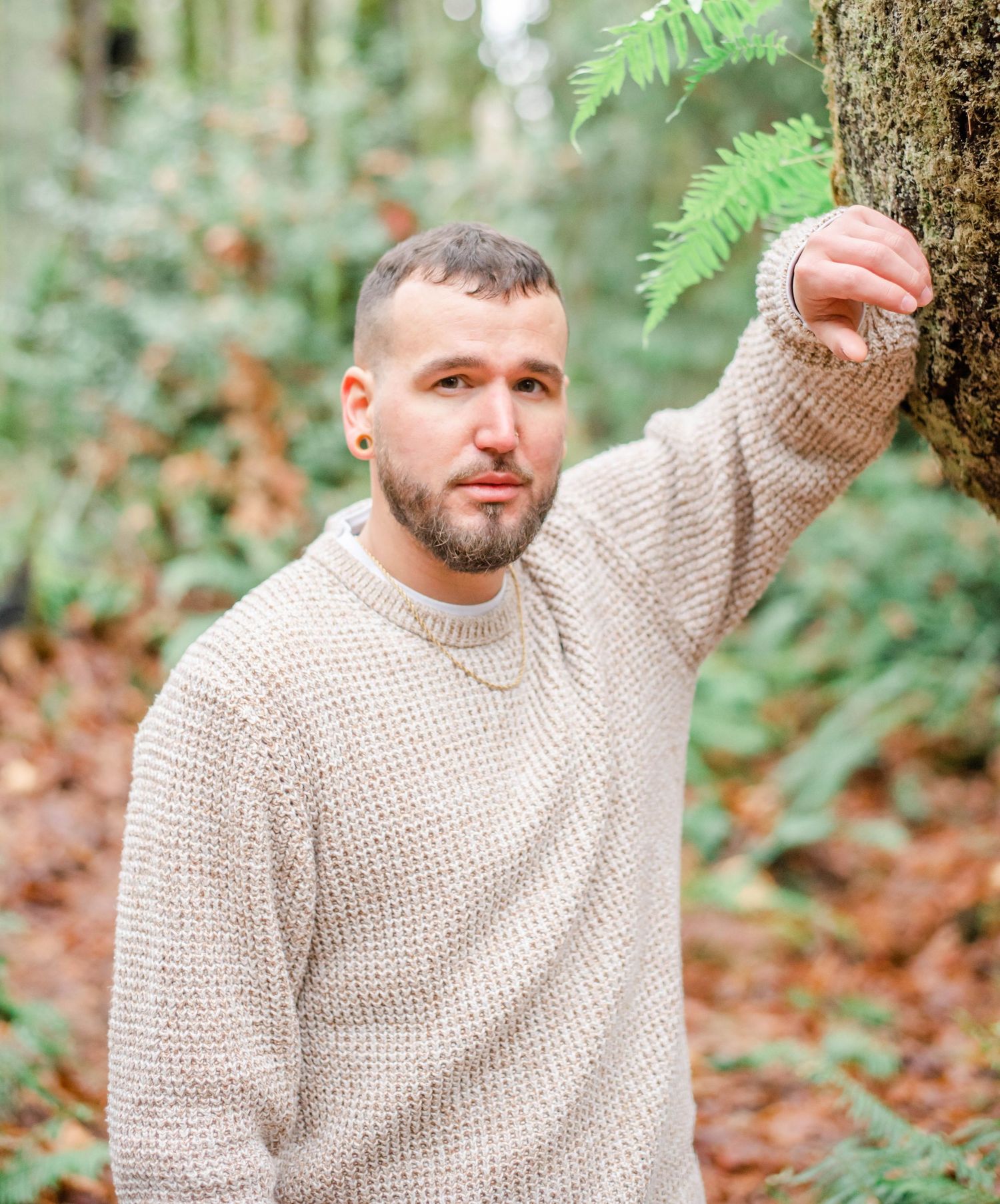 Man in beige sweater poses against tree in forest setting with ferns in background.