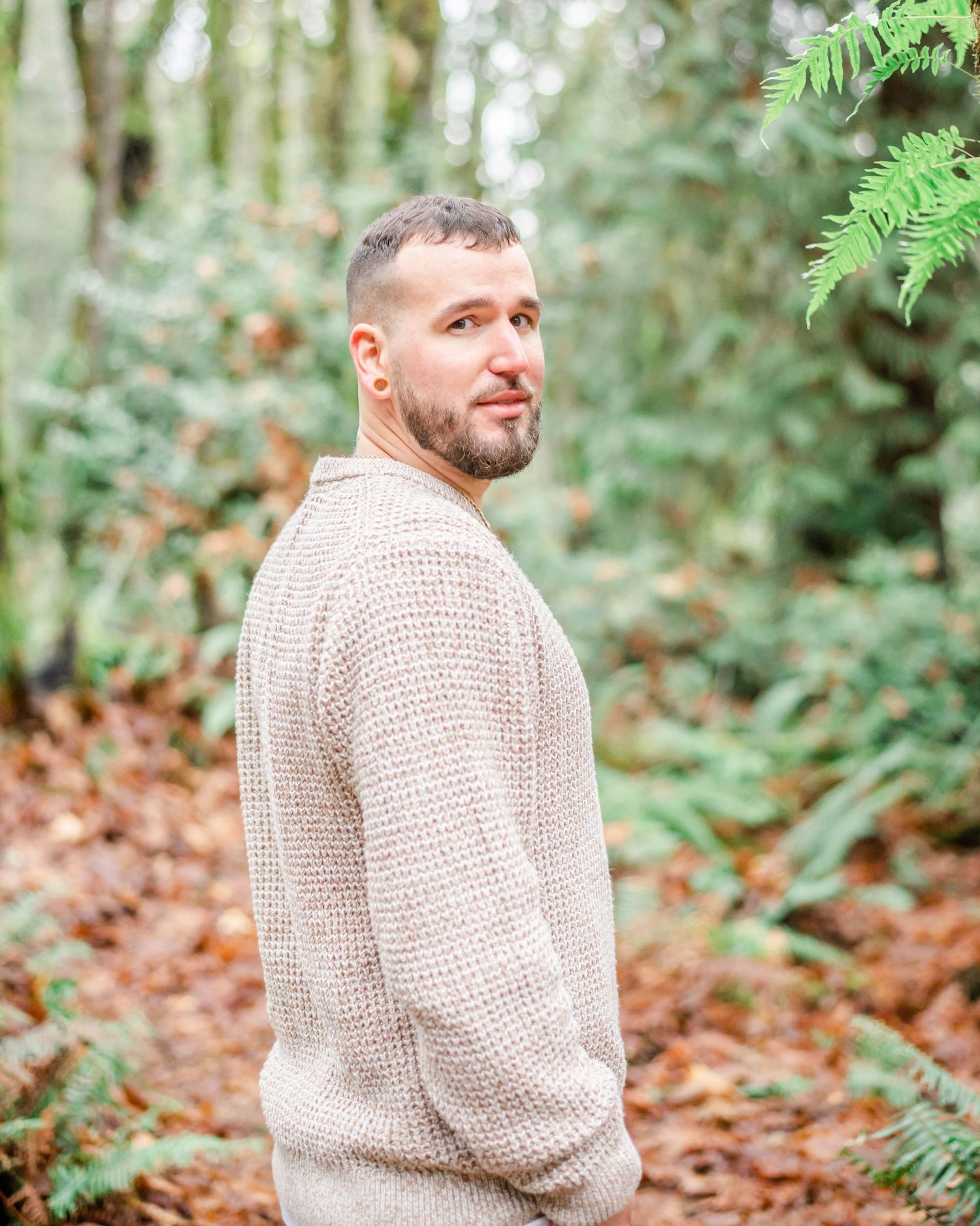 Person wearing a beige knit sweater poses in a forest setting with green ferns and autumn leaves.