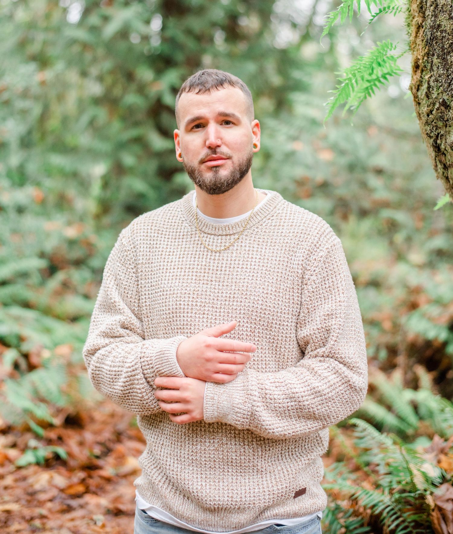 Individual in chunky beige sweater sits outdoors among forest foliage.
