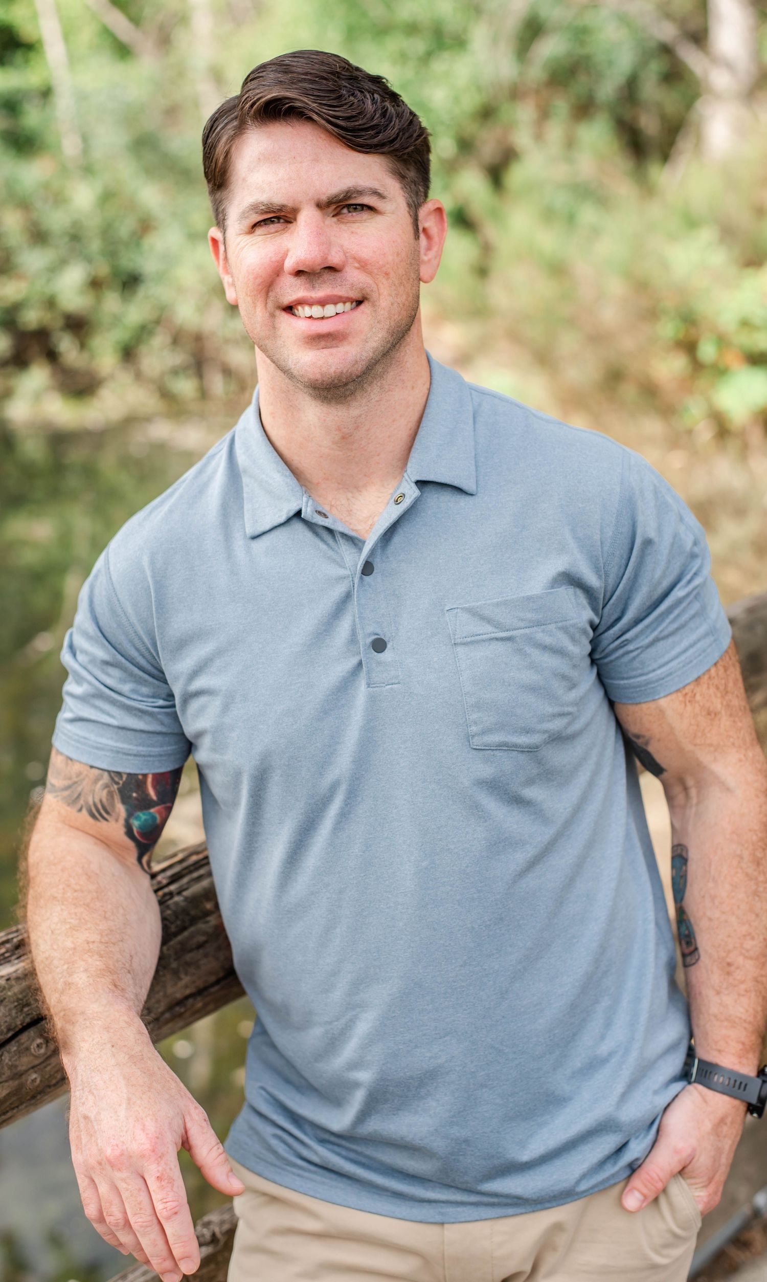 Person in light blue polo and khakis poses casually against wooden railing outdoors.
