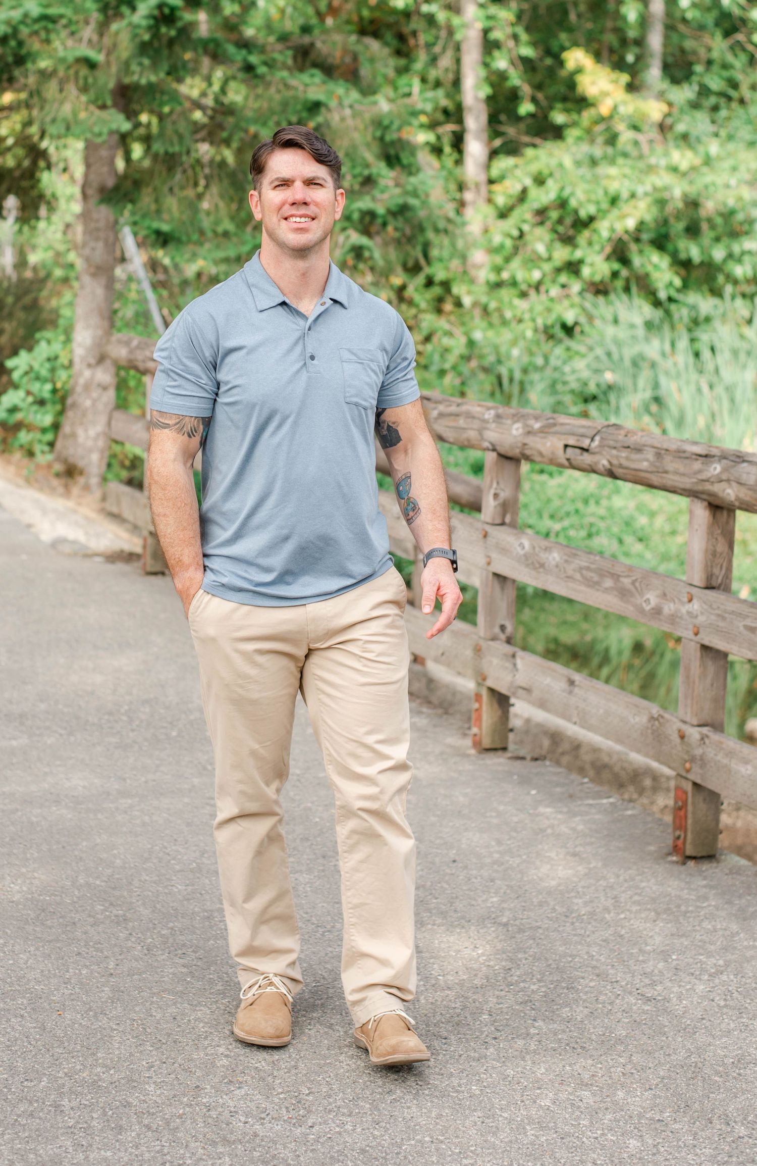Person in gray polo and khakis walking along wooden fence path in park setting.