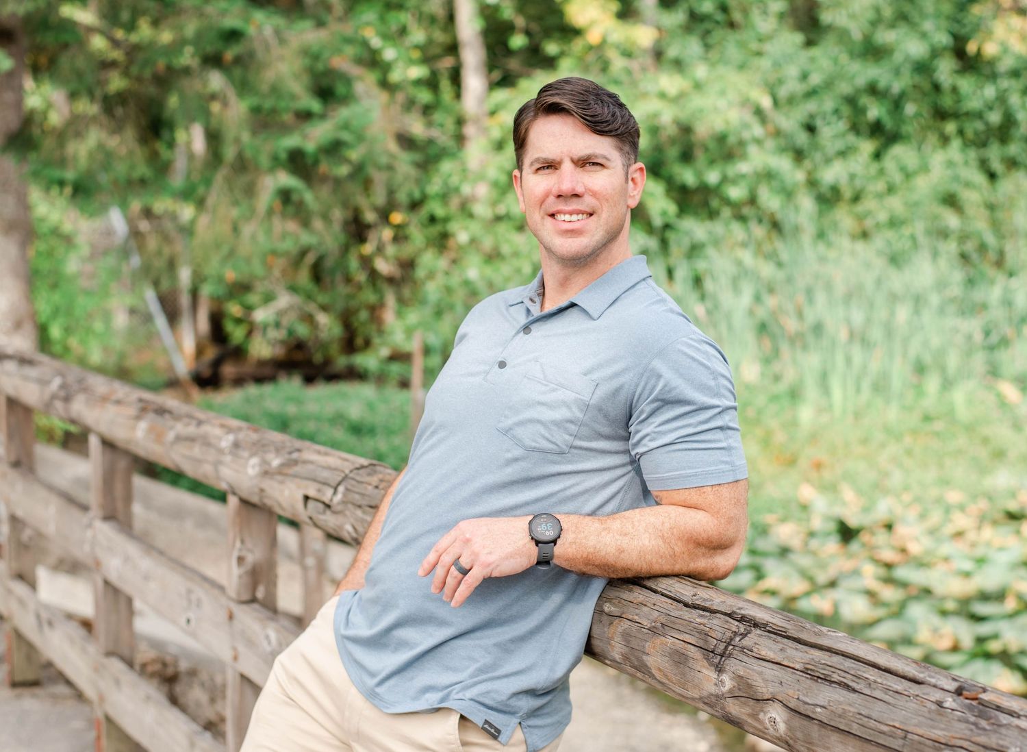 Person wearing blue polo shirt and khaki pants poses casually against wooden fence in outdoor nature setting.
