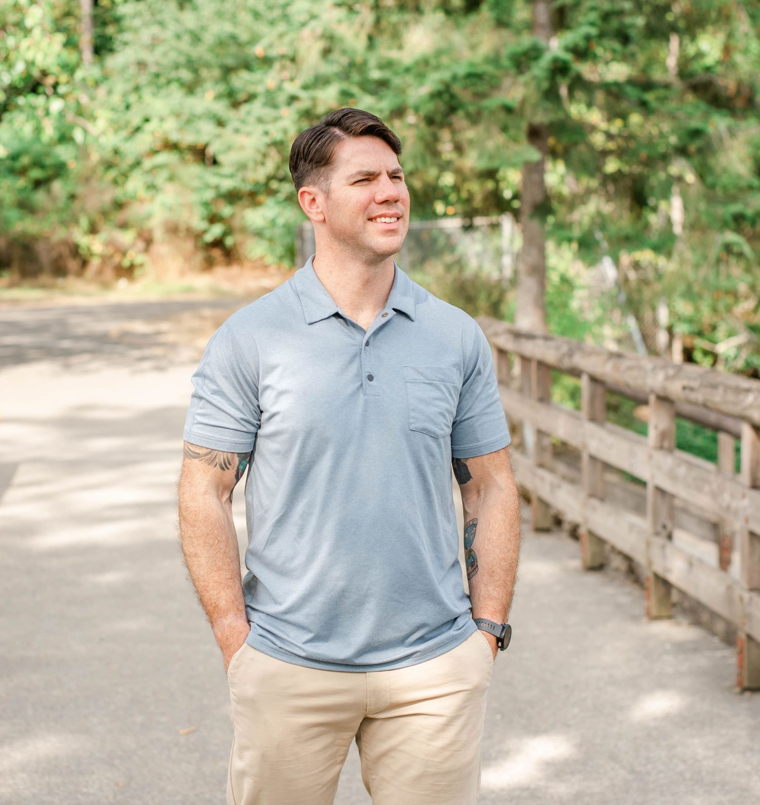 Person in casual attire poses along wooded pathway with wooden fence in sunlit background.