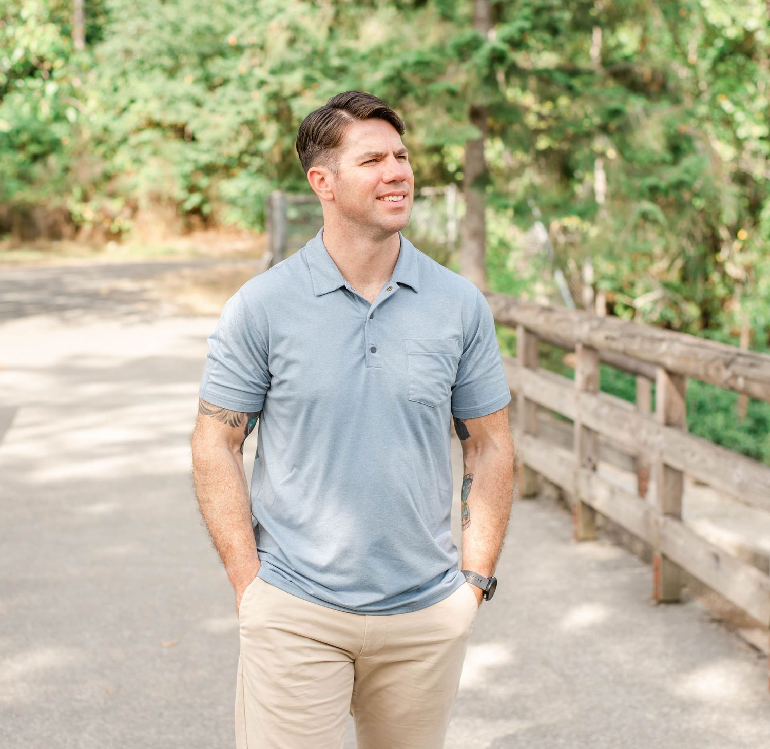 Person in light blue polo shirt and khakis stands on path with rustic wooden railing and greenery.