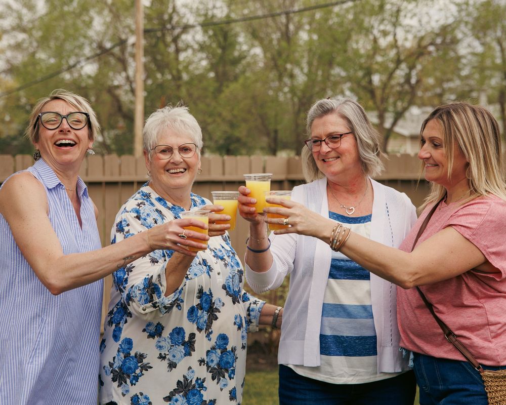 Four women laughing and toasting with orange juice glasses outdoors at a backyard gathering.