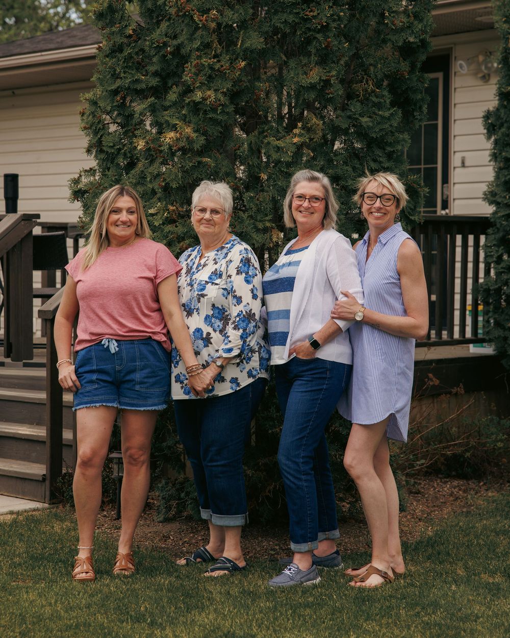 Four women smiling and posing together outdoors in front of a house, wearing casual summer outfits.