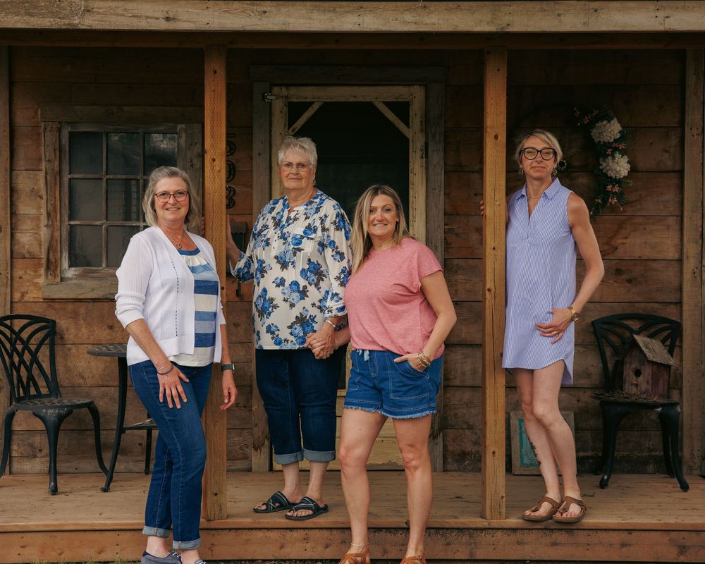 Four women smiling and posing together on a rustic wooden porch of a log cabin.