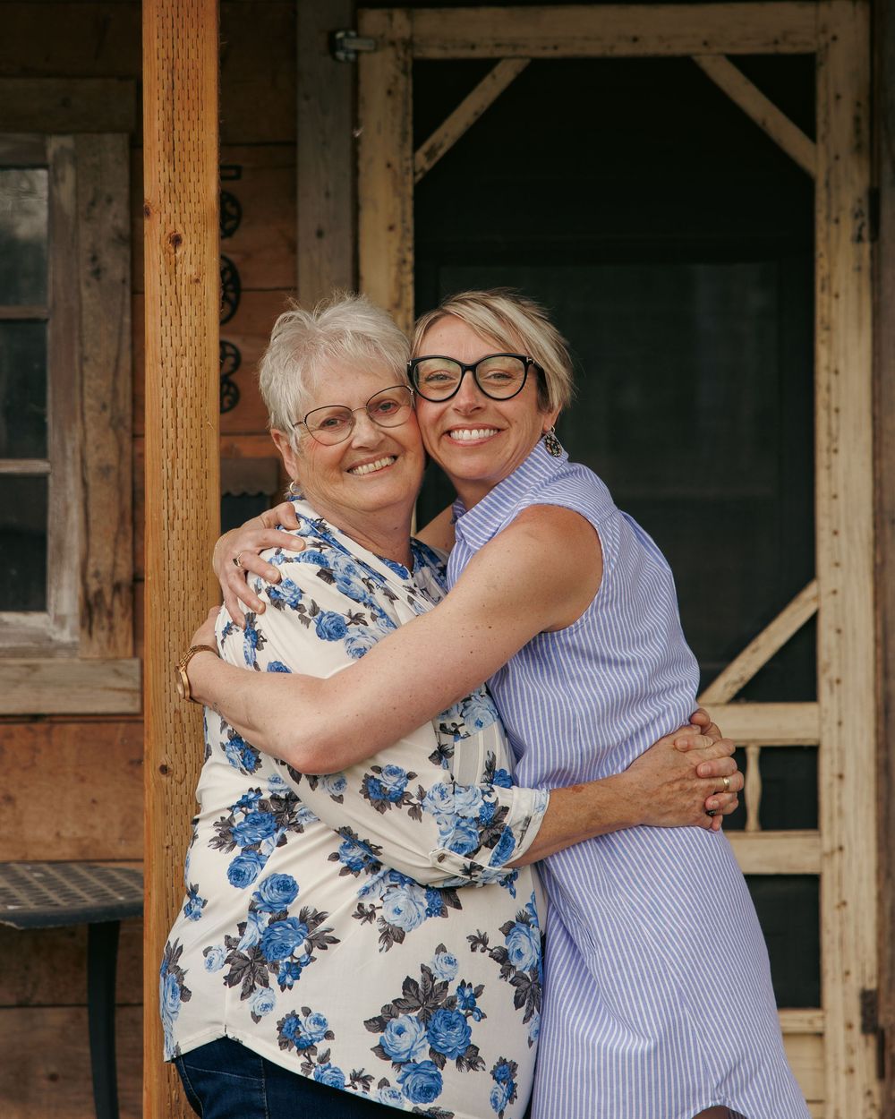 Two women smiling and hugging outdoors near a rustic wooden building, one wearing glasses and a floral shirt.