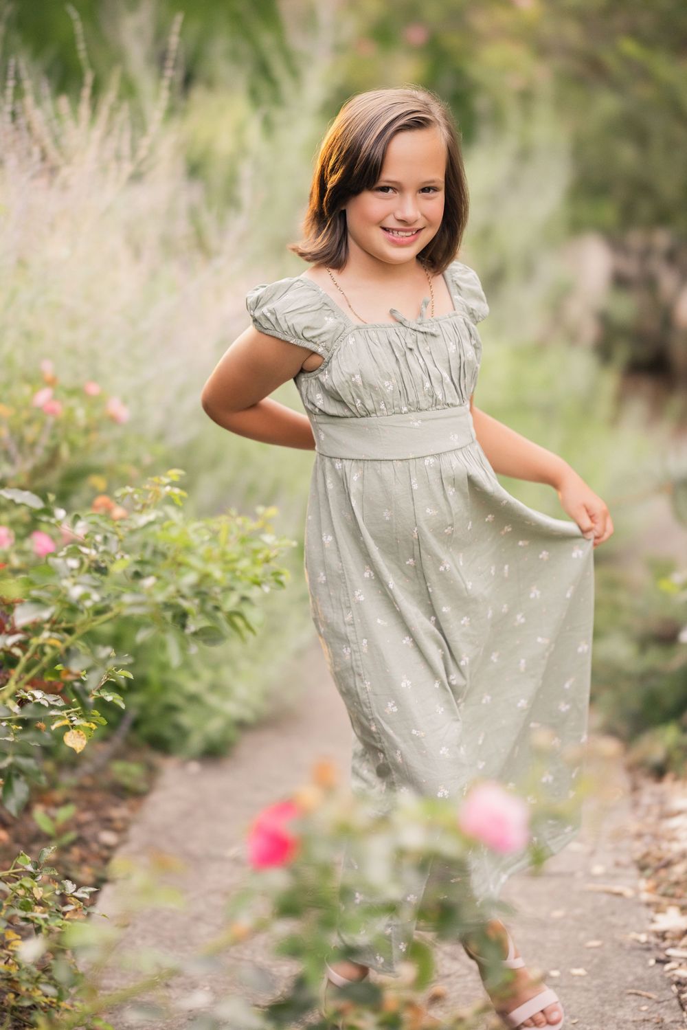 Young person in a light gray dress twirls in a garden path surrounded by flowers.