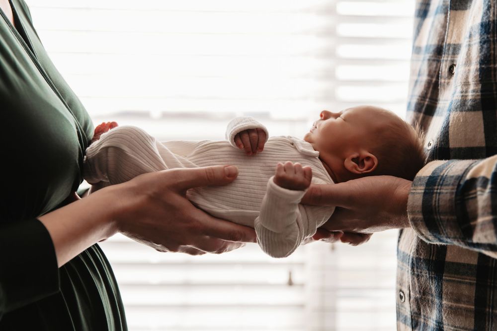 Hands passing a baby bottle in soft window light with gentle shadows.