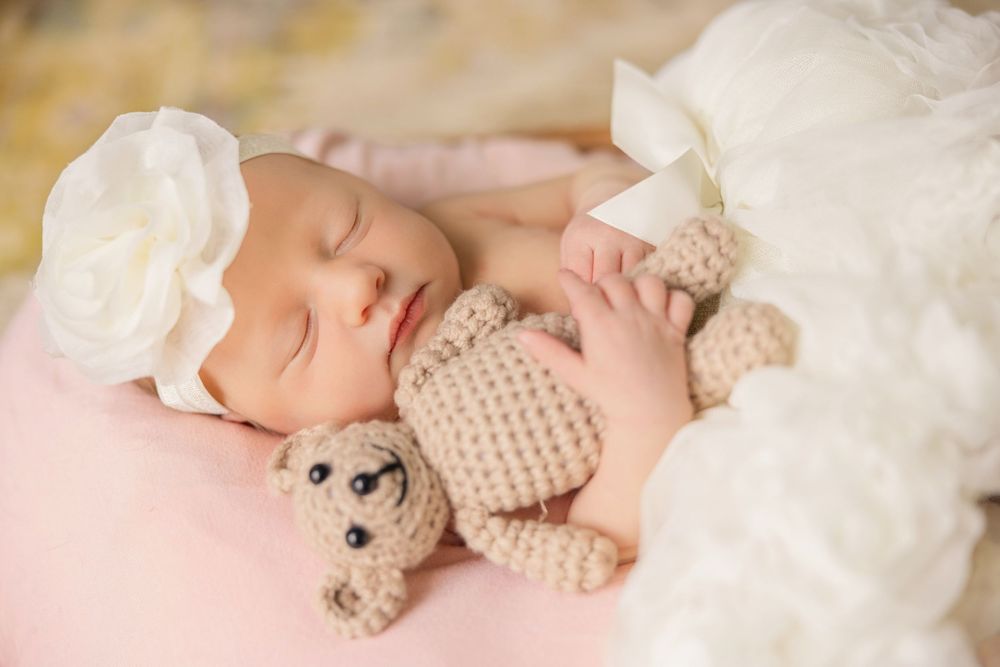 Newborn baby sleeping with small teddy bear on white lace backdrop.