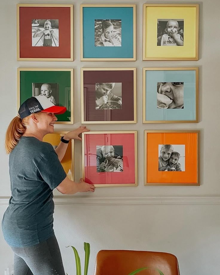 Someone in dark clothing hangs colorful framed photographs in a gallery-style arrangement on a white wall.