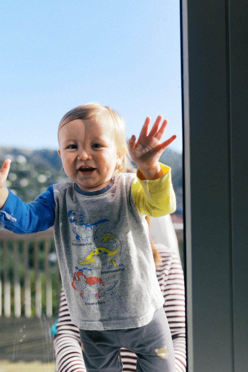 Toddler in striped sleeves and gray sweatshirt making joyful expressions by window.