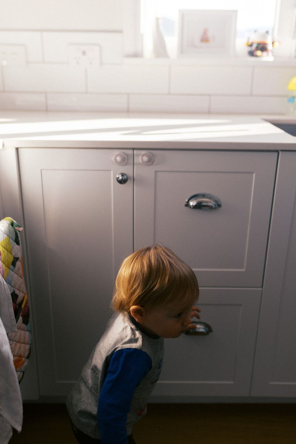 Toddler in blue shirt explores kitchen cabinet storage while crawling along white base cabinets.