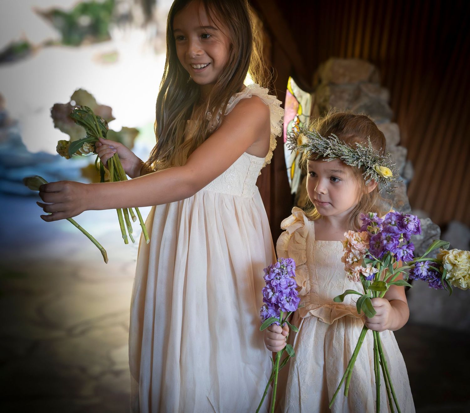Young flower girls in white dresses hold small bouquets with purple flowers during a wedding ceremony at The Sea Ranch Chapel.
